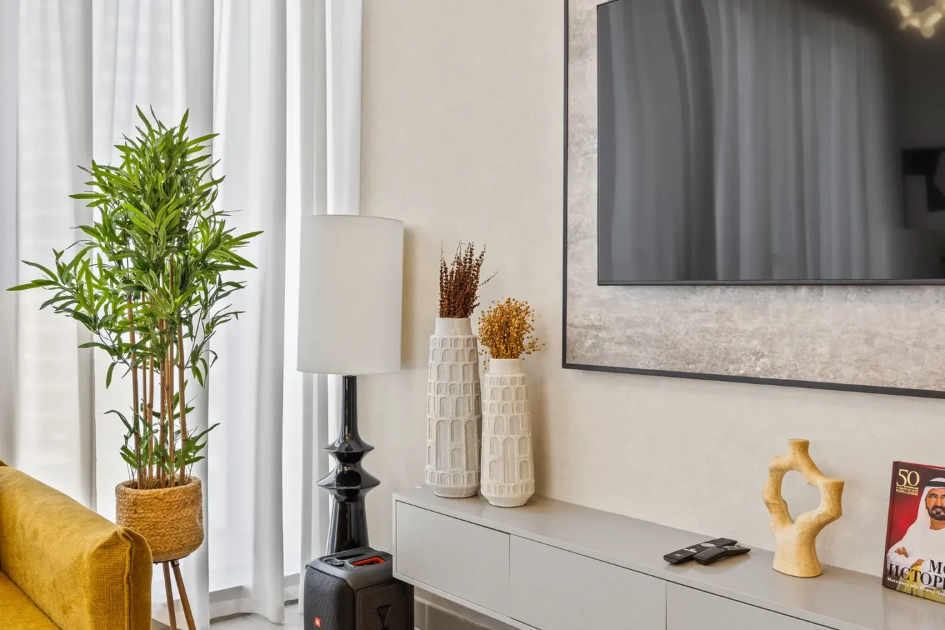 Tall leafy plant in a woven basket beside a white-curtained window; a modern black-based lamp and two textured vases sit on a gray media console under a wall-mounted TV.