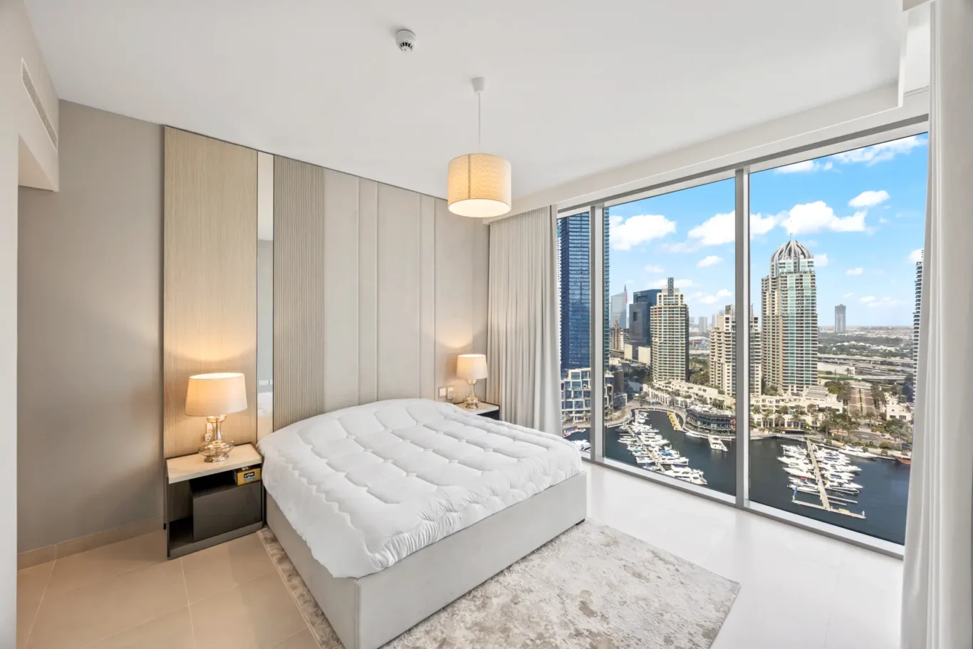 Modern bedroom with a tufted white bed, mirrored headboard panels, and floor-to-ceiling windows overlooking a marina and city skyline.