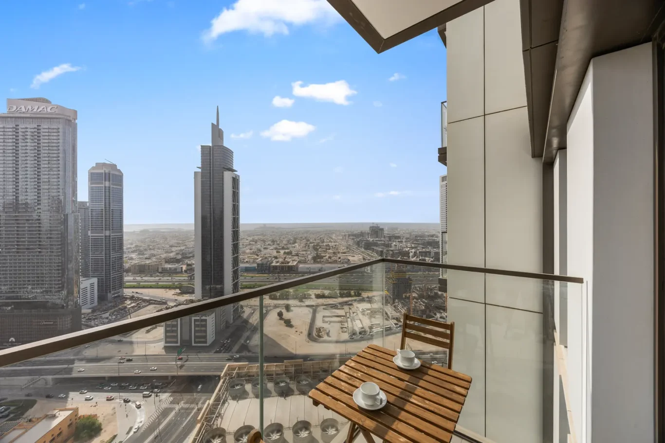 Balcony with a wooden table and two cups overlooking a city with tall modern skyscrapers and a blue sky in the distance.