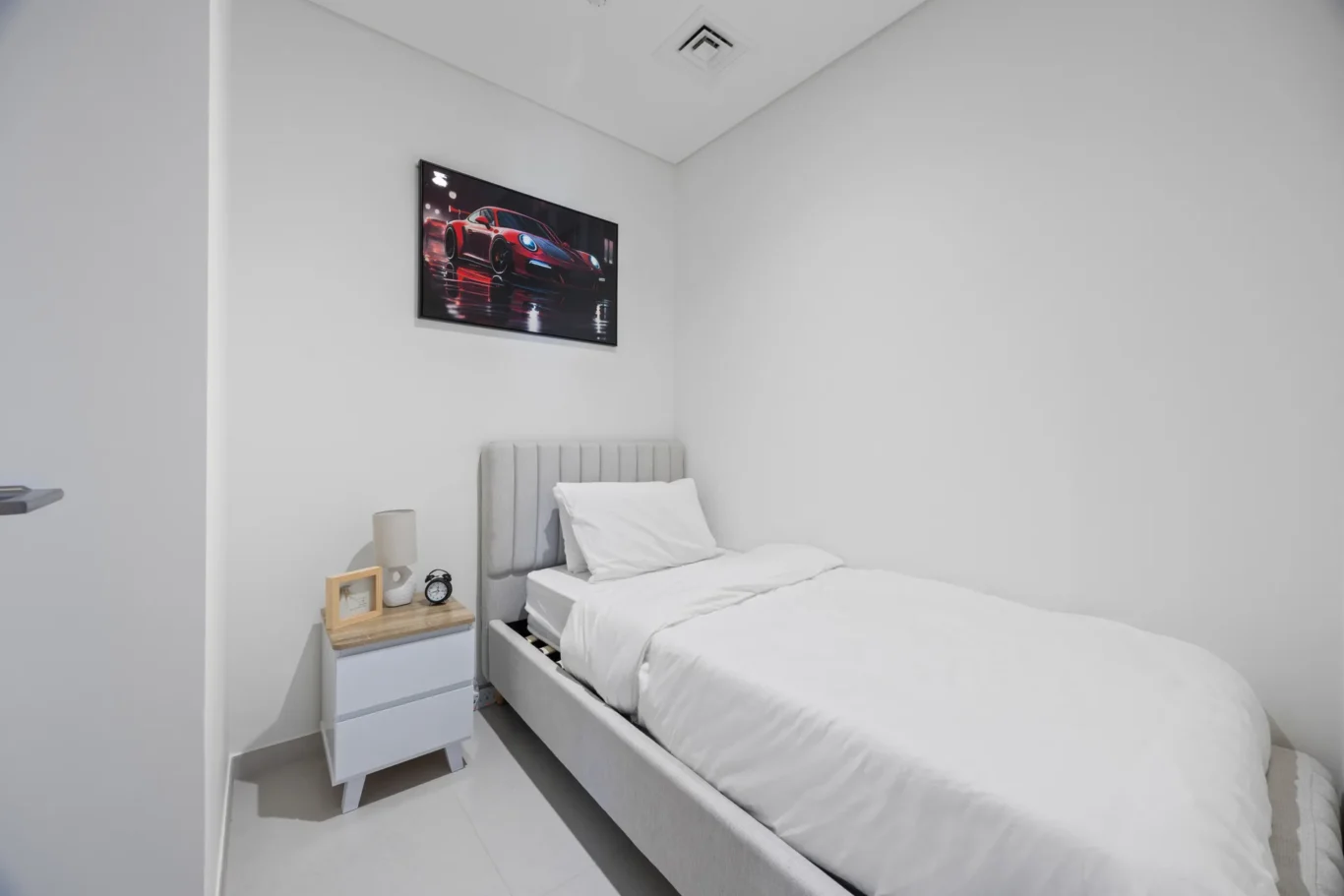 Small white bedroom with a gray upholstered bed, white bedding, a wooden nightstand, alarm clock and a framed car poster on the wall.