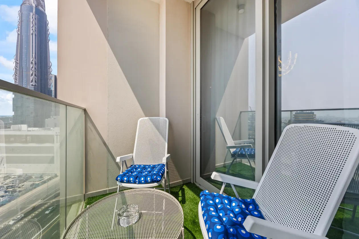 Balcony seating area with white mesh chairs and blue patterned cushions on artificial grass, overlooking a city skyline.