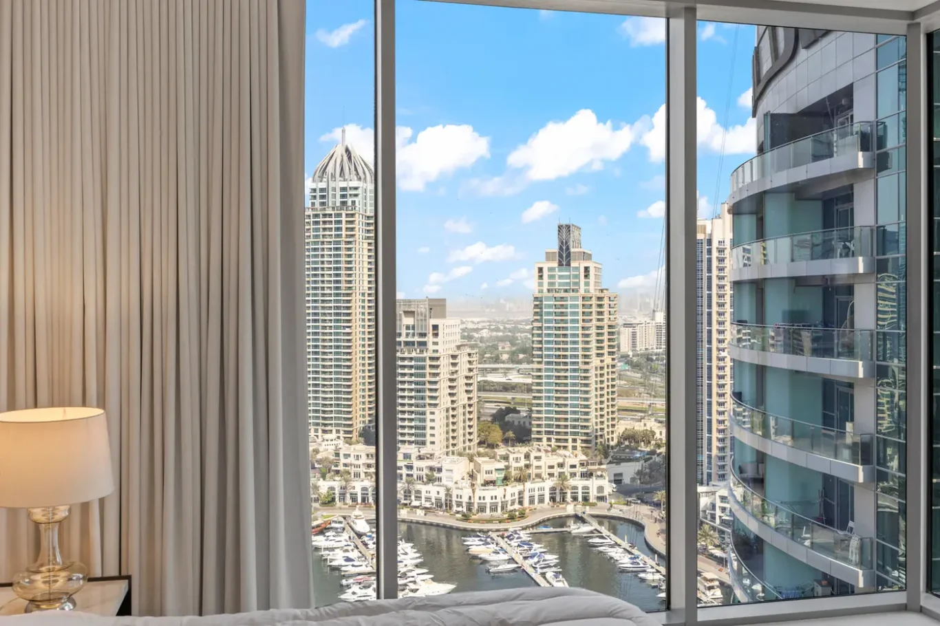 Hotel room with a floor-to-ceiling window overlooking a marina and city skyline on a sunny day, lamp and curtains visible inside.