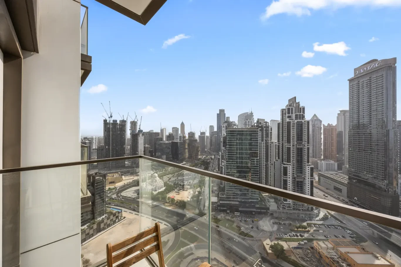 City skyline viewed from a balcony with glass railing, tall modern skyscrapers and construction cranes under a blue sky.