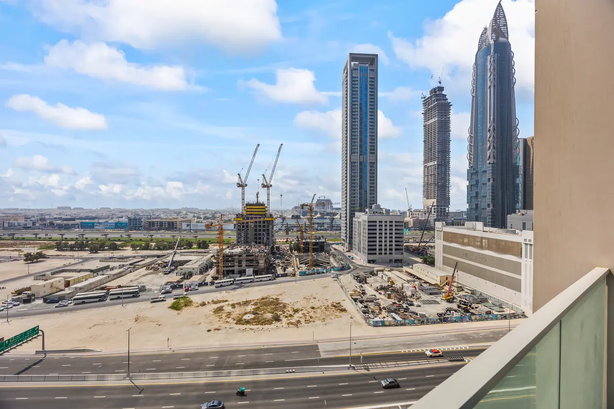 Construction site in a modern city with cranes, mid-rise and high-rise buildings, and a multi-lane road in the foreground viewed from a balcony/terrace edge.