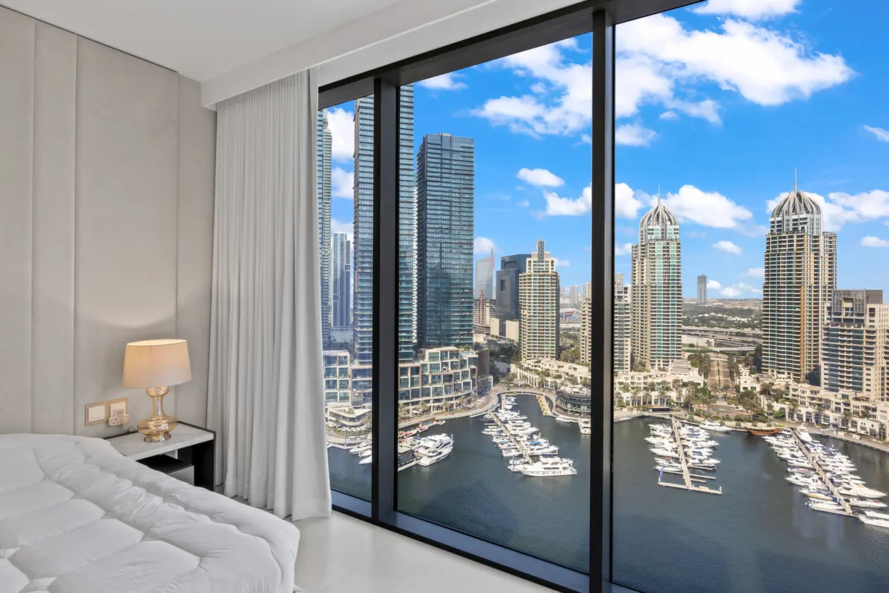 Bedroom with floor-to-ceiling glass window showing a marina and skyline of tall buildings; white bedding and a bedside lamp visible to the left.