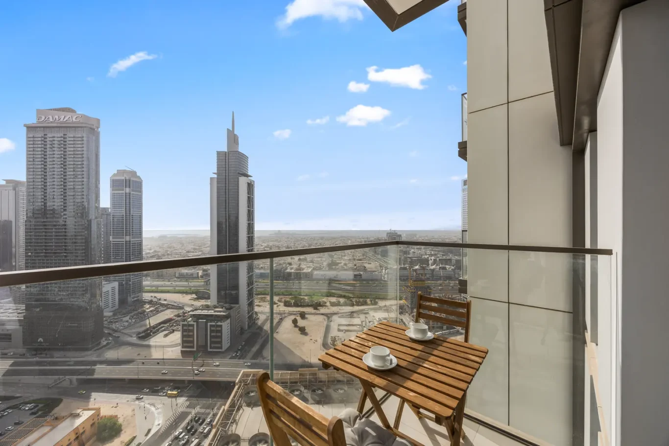 Balcony table with two cups overlooking a city skyline of tall glass buildings and clear blue sky.