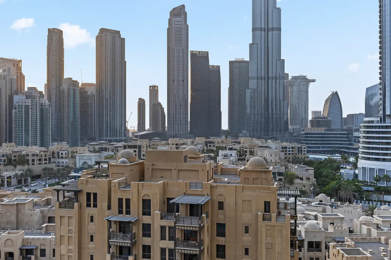 Dubai-style city skyline with beige low-rise buildings in the foreground and tall glass skyscrapers against a blue sky.