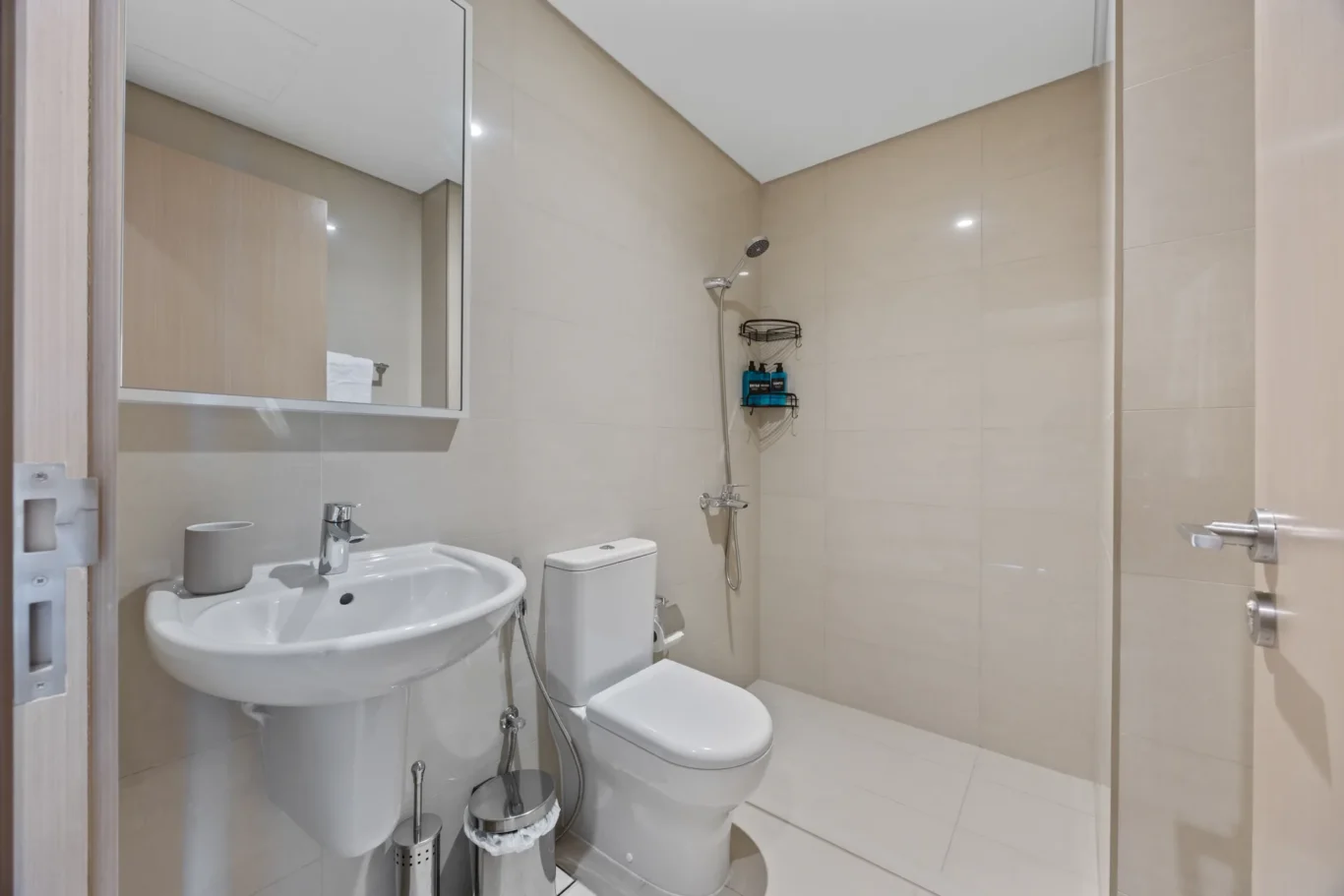 Modern beige-tiled bathroom with a white pedestal sink, toilet, and a glass-free shower; wall mirror cabinet reflects part of the room.