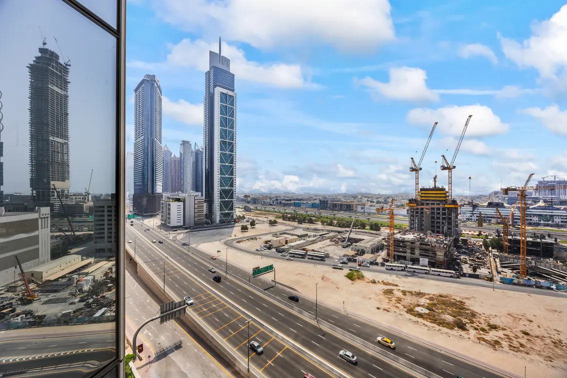 Skyline of a modern city with tall glass towers and active construction cranes, seen from a high-rise window over a busy freeway of multiple lanes.