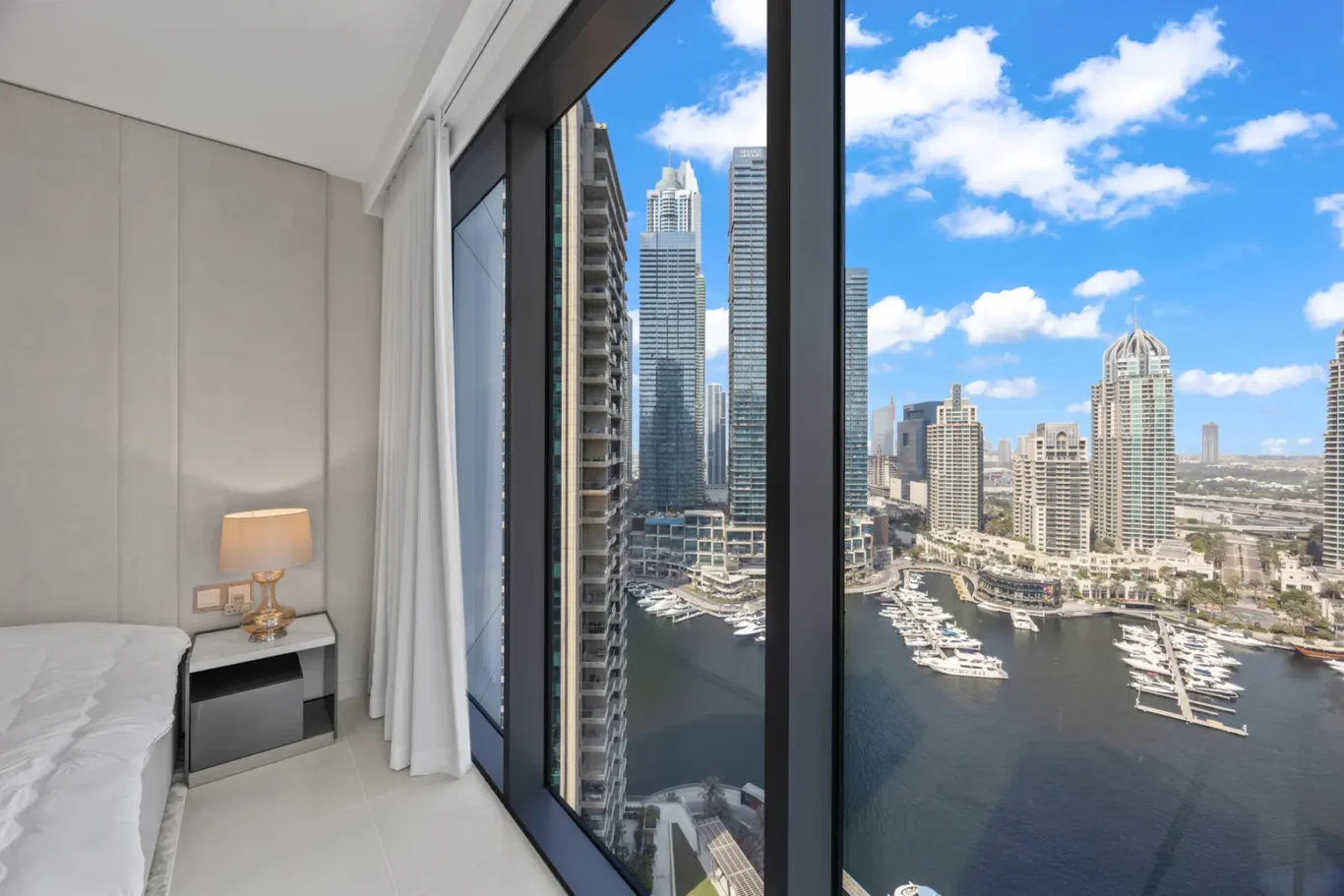 Bedroom with a large window view of a marina and city skyline, lamp and nightstand beside the bed inside a modern room.