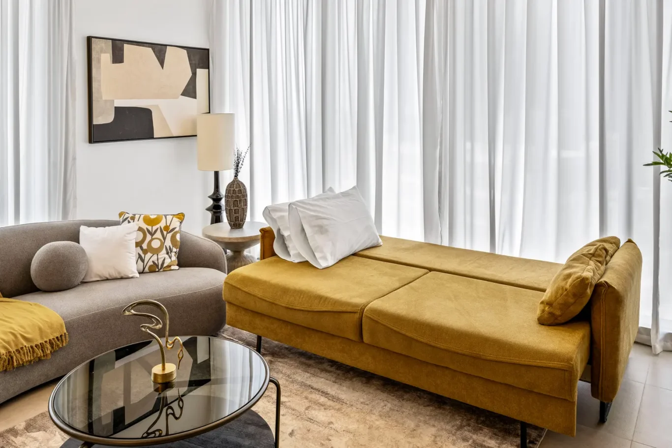 Modern living room with a mustard chaise lounge, a gray sectional, and white drapes; abstract artwork on the wall and a glass coffee table in front.