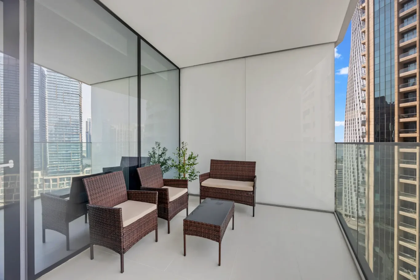 Modern apartment balcony with brown wicker chairs and a matching table, glass railing, overlooking city skyscrapers.