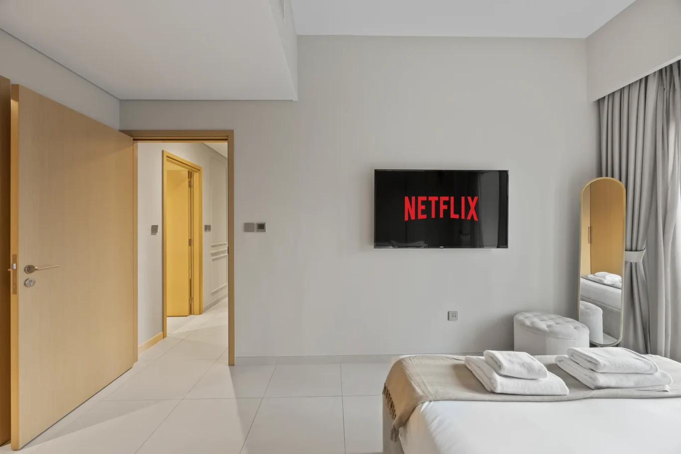 Hotel bedroom with a wall-mounted TV showing Netflix, two neatly made beds with folded towels, and a full-length mirror by the window curtains.