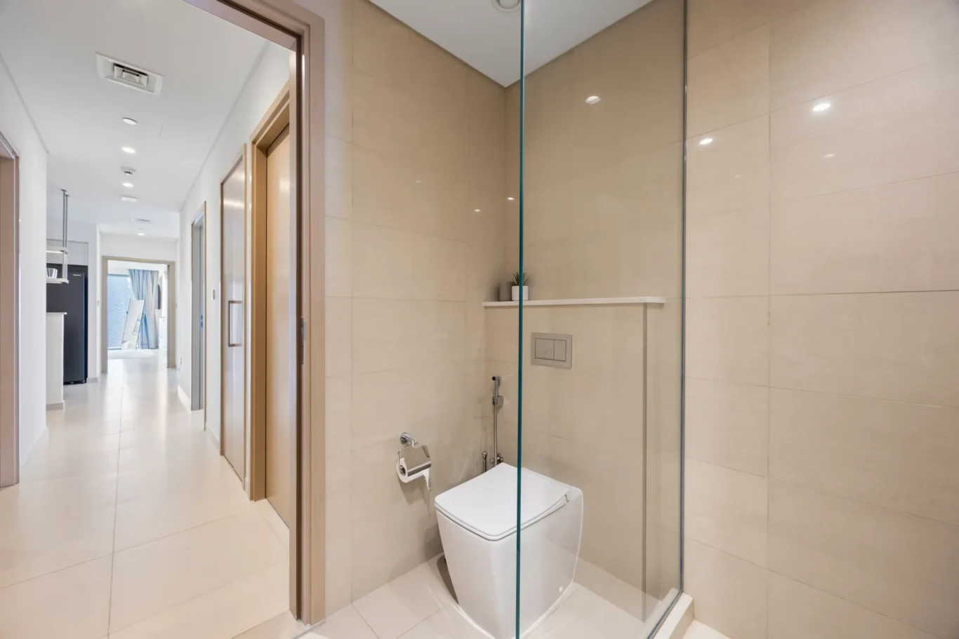 Modern bathroom with a glass shower enclosure and a wall-hung toilet; beige tiled walls and a bright hallway beyond.