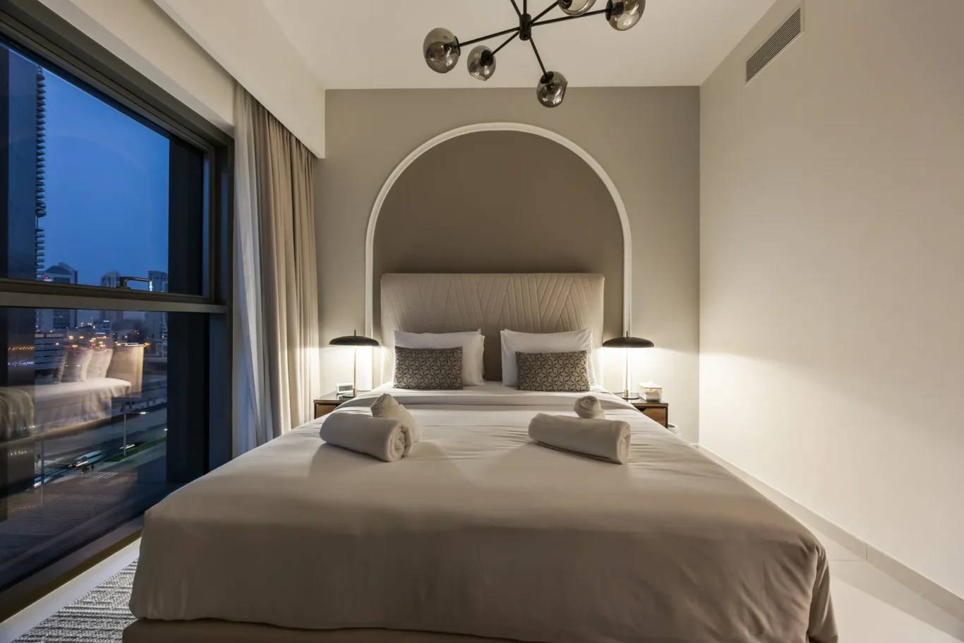 Hotel bedroom with a large bed and a curved arch headboard, two decorative pillows, and city lights visible through a window at dusk/night.
