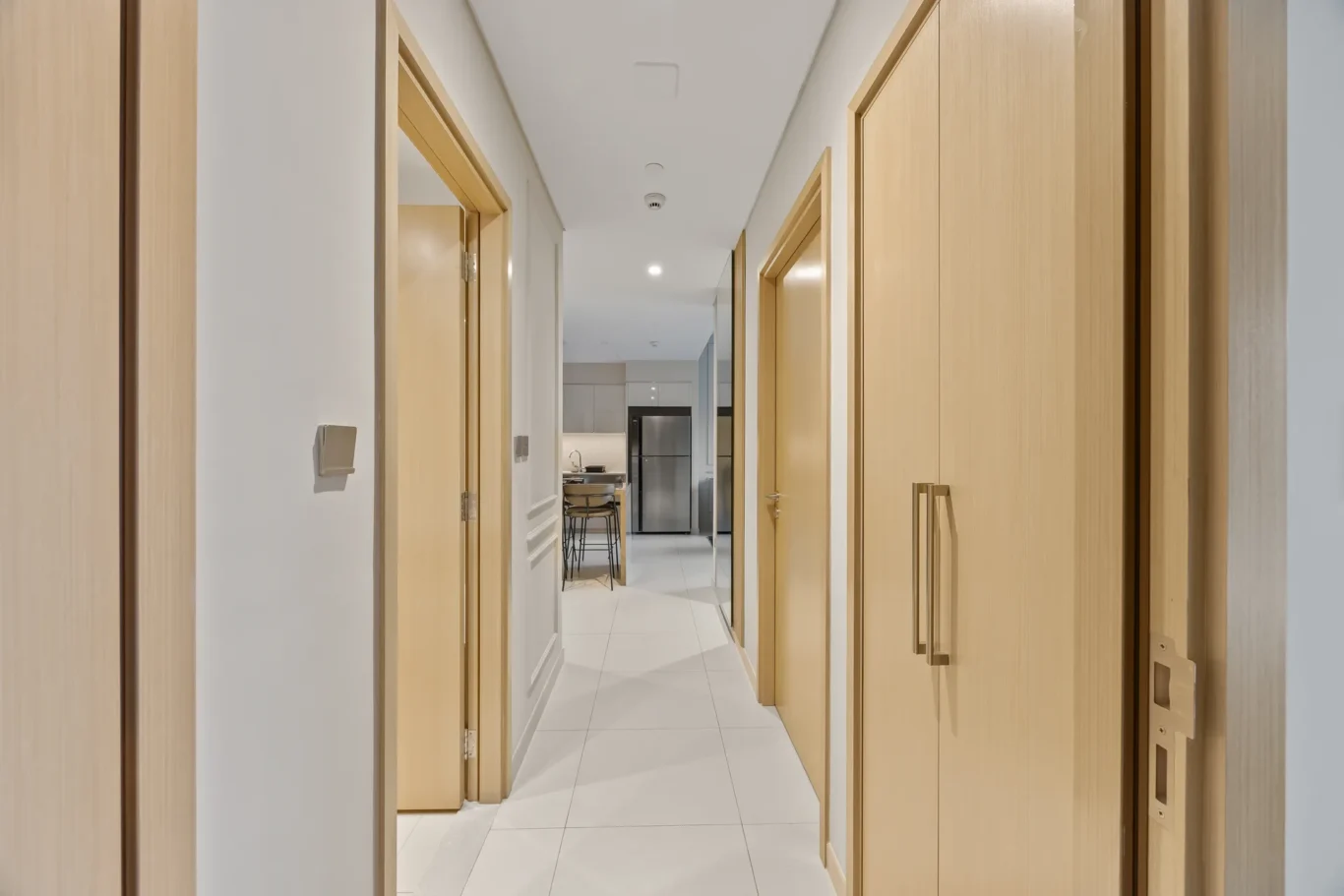 Bright hallway with light wood doors on both sides leading to a modern kitchen at the far end, stainless steel fridge visible.