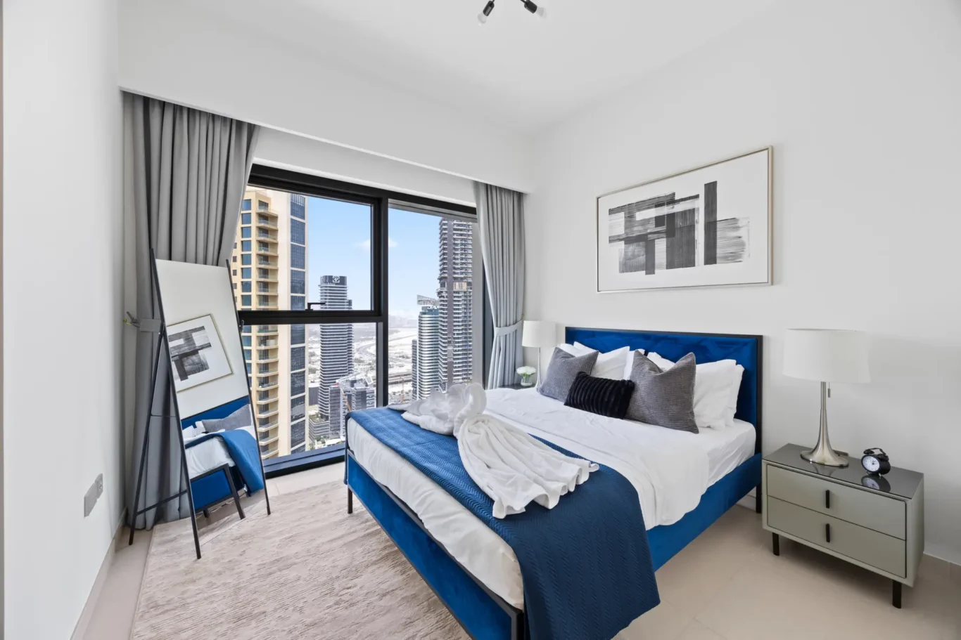 Modern bedroom with a blue upholstered bed, white and blue bedding, gray curtains, and a city view through a large window, sunny and bright.