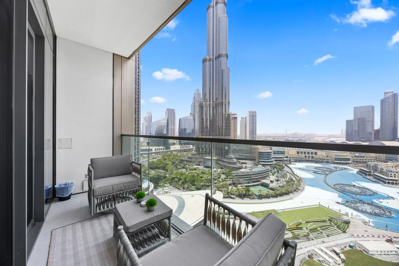 Balconity seating area with gray cushions, glass railing, and a skyline featuring the Burj Khalifa above a curved resort pool.