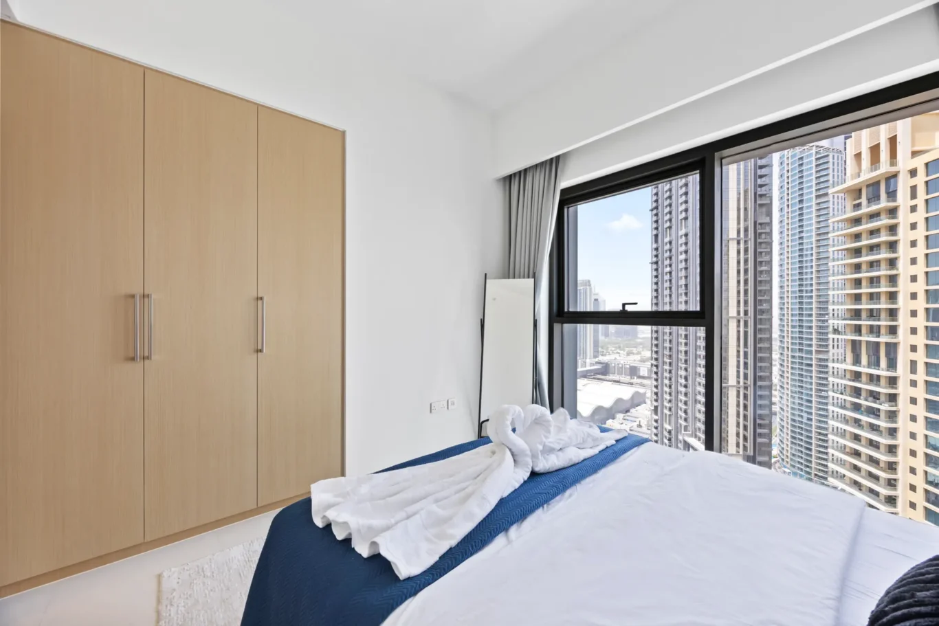 Bedroom with a tall light-wood wardrobe on the left and a large window showing city skyscrapers; bed with white sheets and a blue throw.
