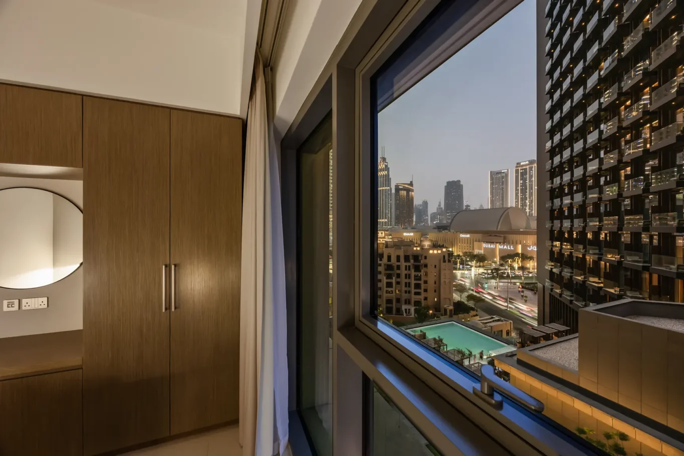 Hotel room interior with wooden wardrobes and a round mirror, open window showing Dubai’s illuminated skyline at night and a pool below.