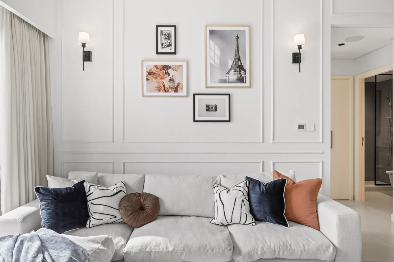 Living room scene with a light gray sofa and decorative cushions in front of paneled white walls and a collage of framed art above.