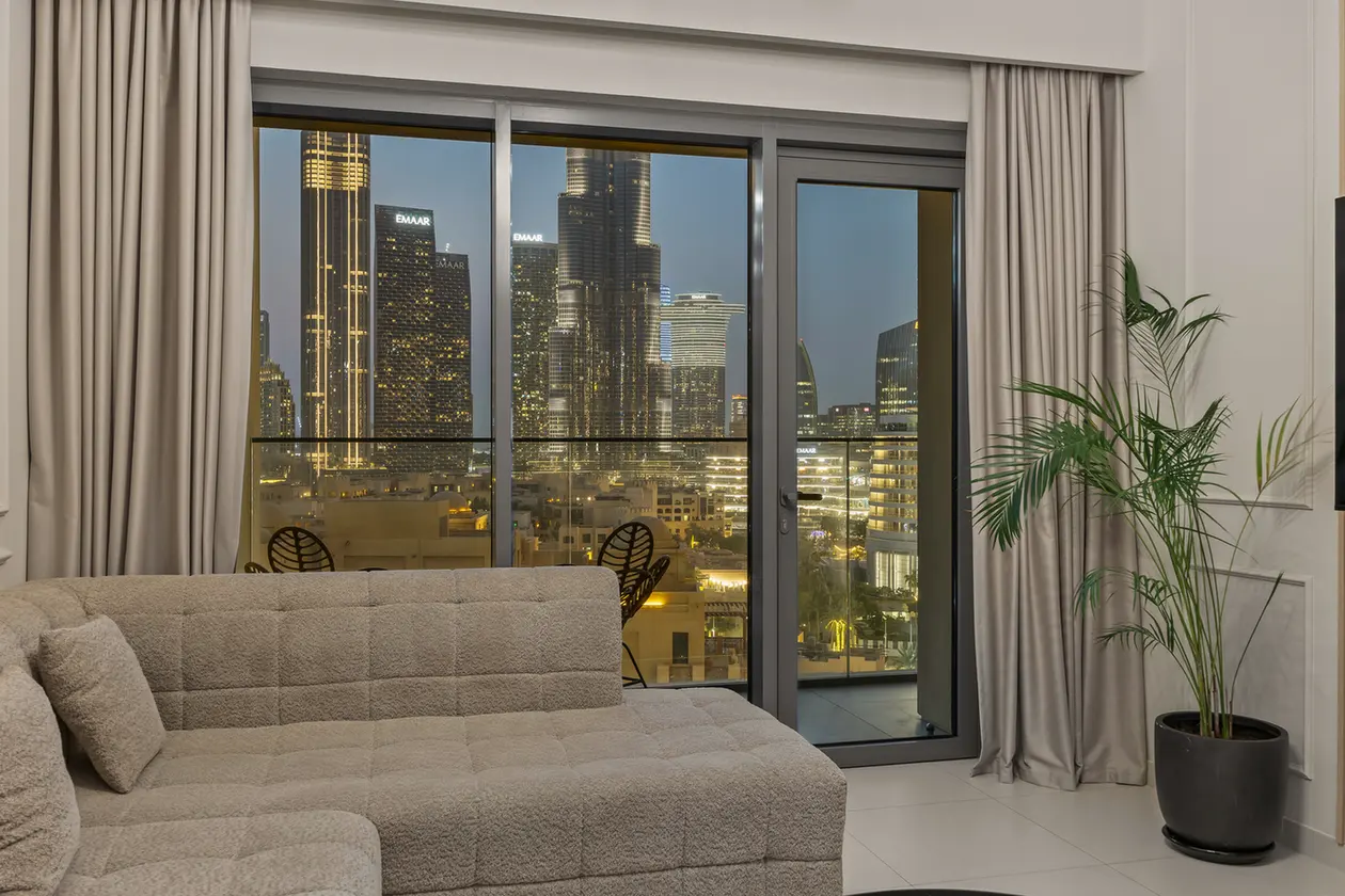 Living room with a beige tufted sofa and large glass doors showing a city skyline at night.