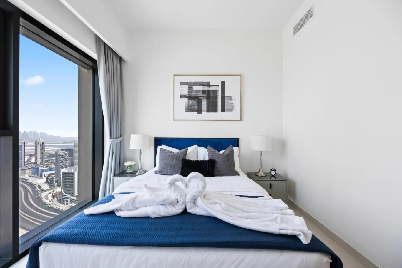 Modern hotel bedroom with a large window showing the city skyline, a blue upholstered headboard, white bedding, and an abstract framed artwork above the bed.