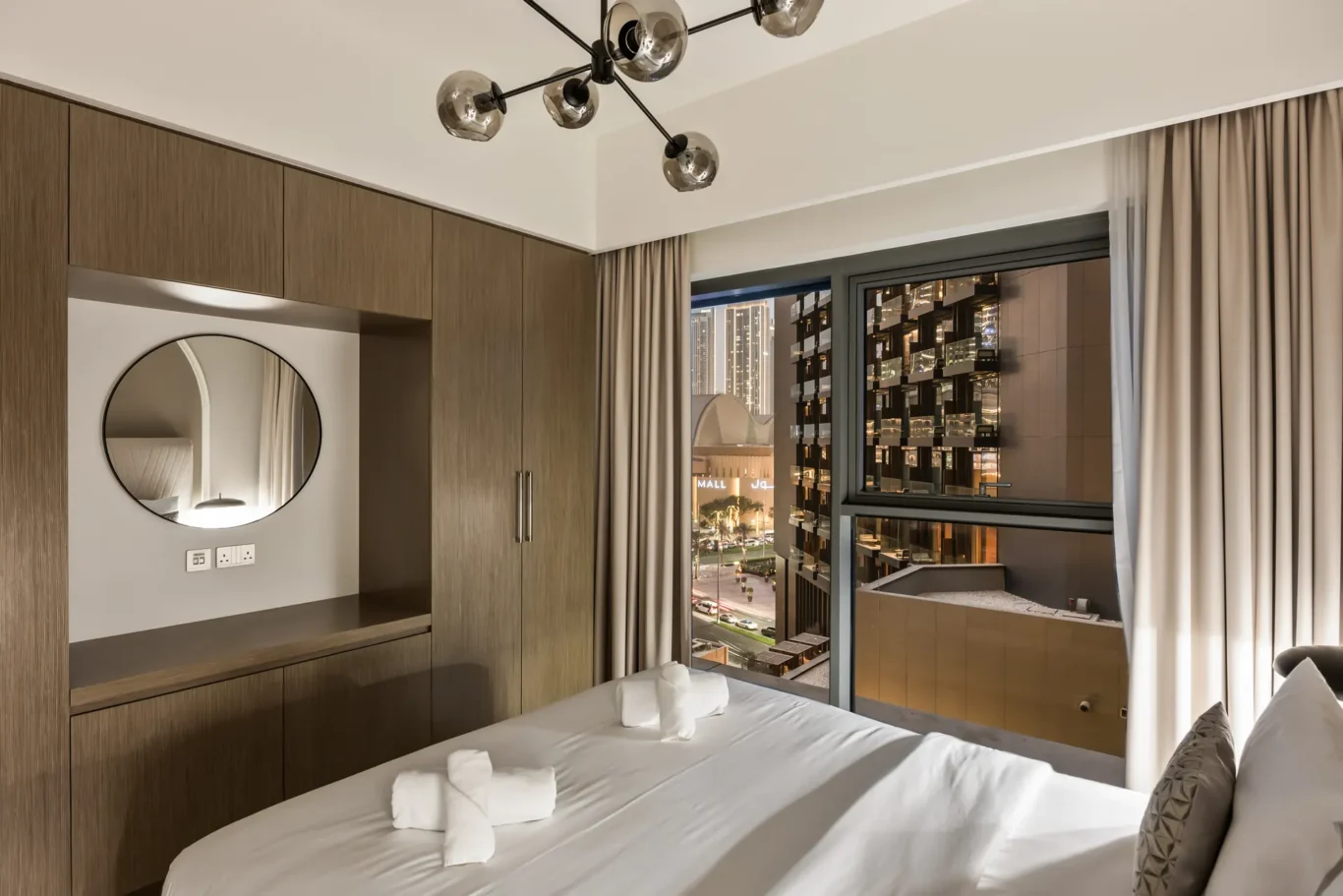 Hotel bedroom featuring built-in wooden wardrobes, a circular mirror, and a tidy bed with rolled towels; large window reveals city lights outside.