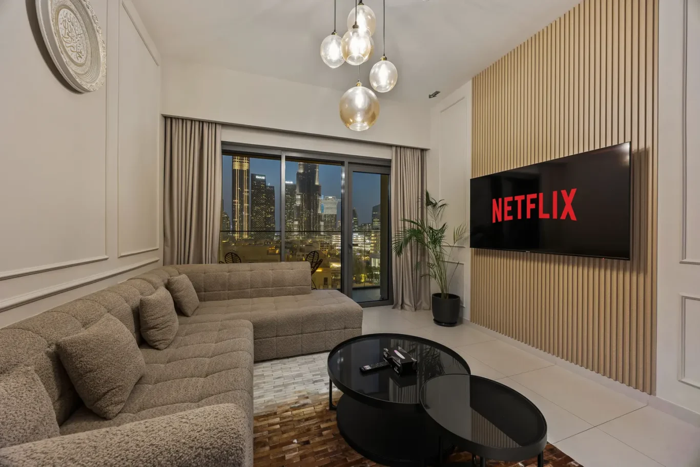 Modern living room with an L-shaped beige sofa, glass coffee tables, and a wall-mounted Netflix screen on a wooden slat wall; city skyline visible through large windows. Thinks to note: pendant globe lights above and a potted plant nearby.