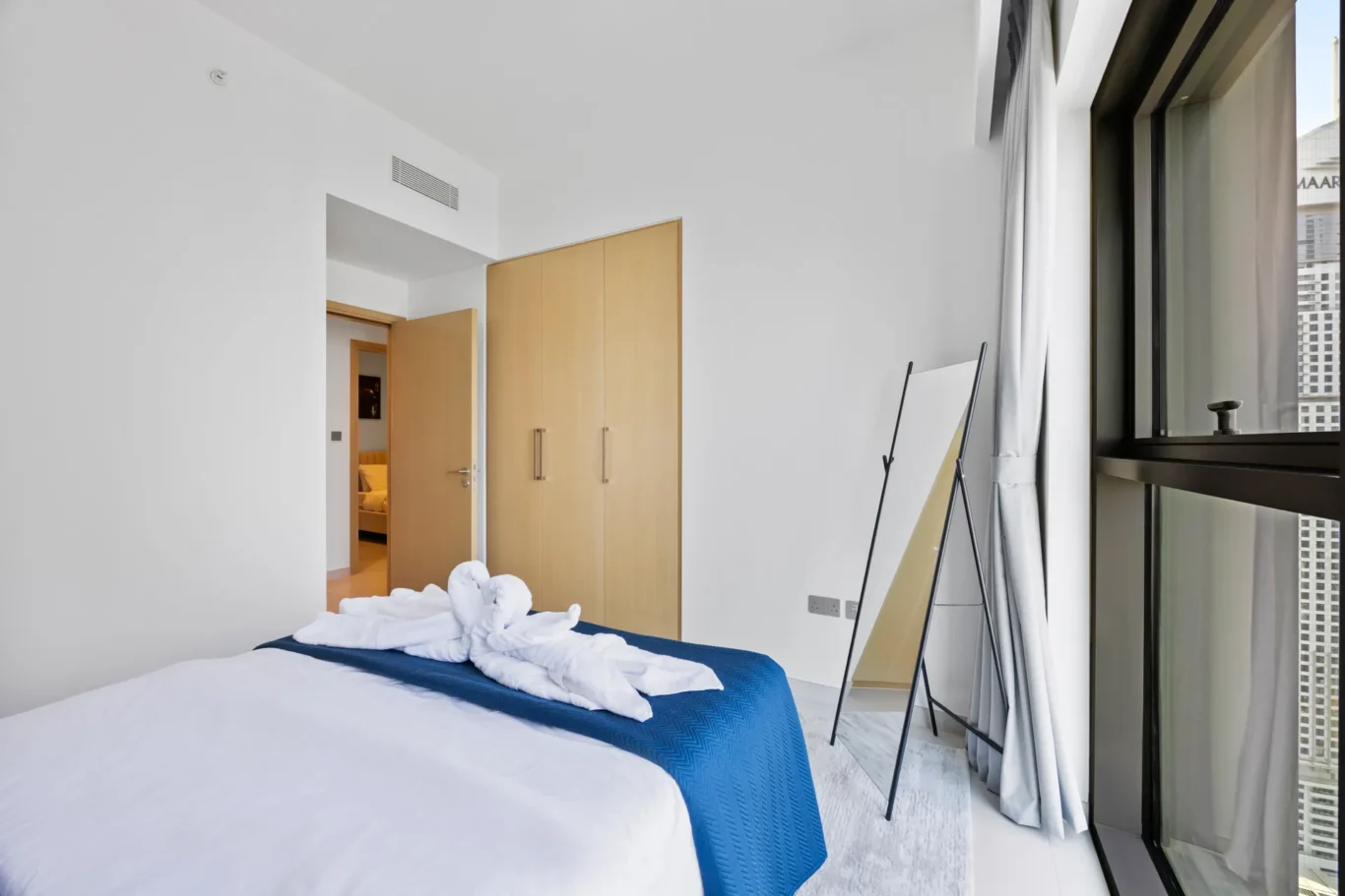 Modern hotel bedroom with a white bed, blue throw, and towel art near the foot of the bed; large window with curtains and a city view on the right.