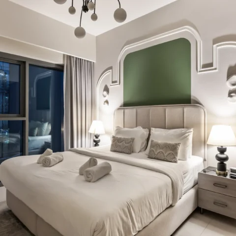 Hotel bedroom with a large upholstered bed, matching nightstands, and decorative wall alcove; window with curtains to the left on a night view backdrop.