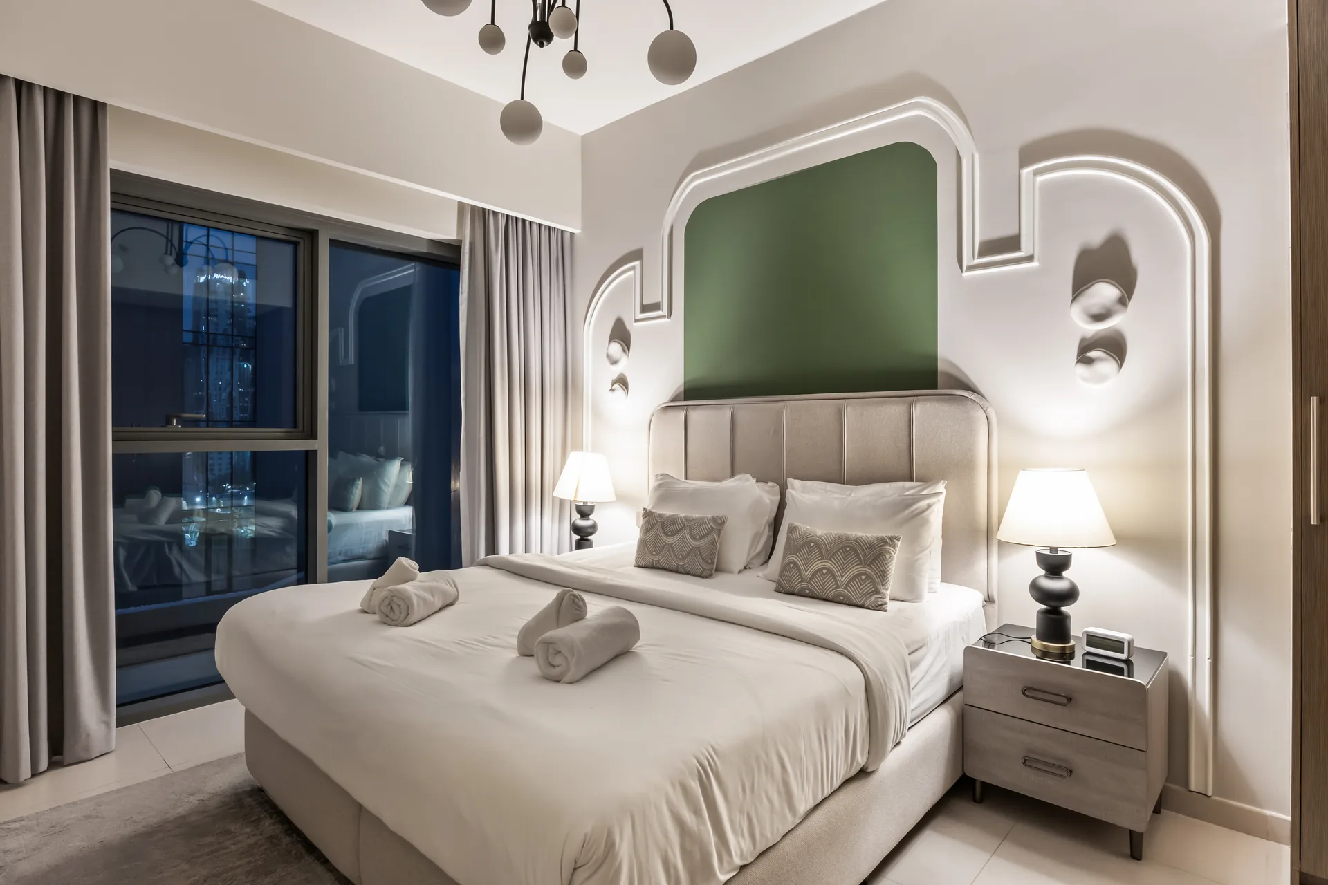 Hotel bedroom with a large upholstered bed, matching nightstands, and decorative wall alcove; window with curtains to the left on a night view backdrop.