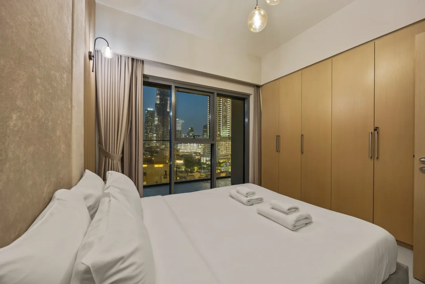 Hotel room with a large bed in foreground, white linens and folded towels, and a city skyline visible through floor-to-ceiling windows with beige curtains and built-in wooden wardrobes along the right side