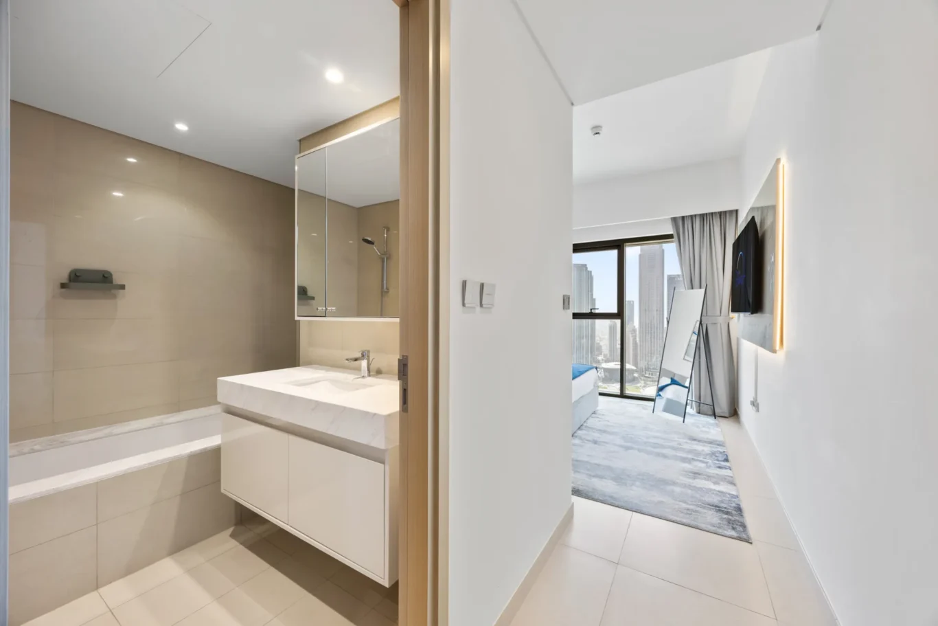Open doorway from a beige-tiled bathroom into a bright hotel suite with a city skyline view.