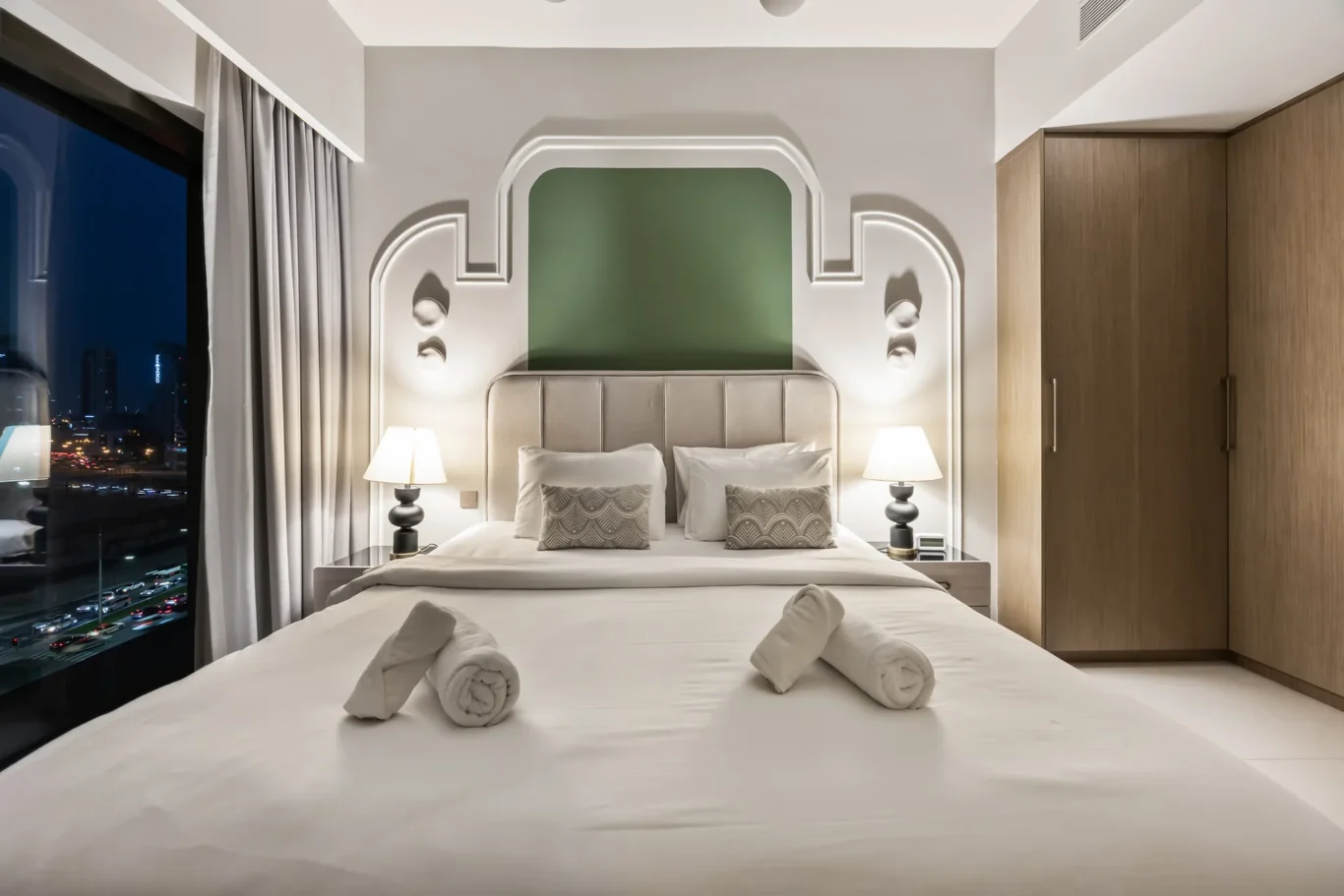 Hotel room with a large bed, tufted headboard, two bedside lamps, and towels rolled on the sheets; city lights visible through a window to the left.