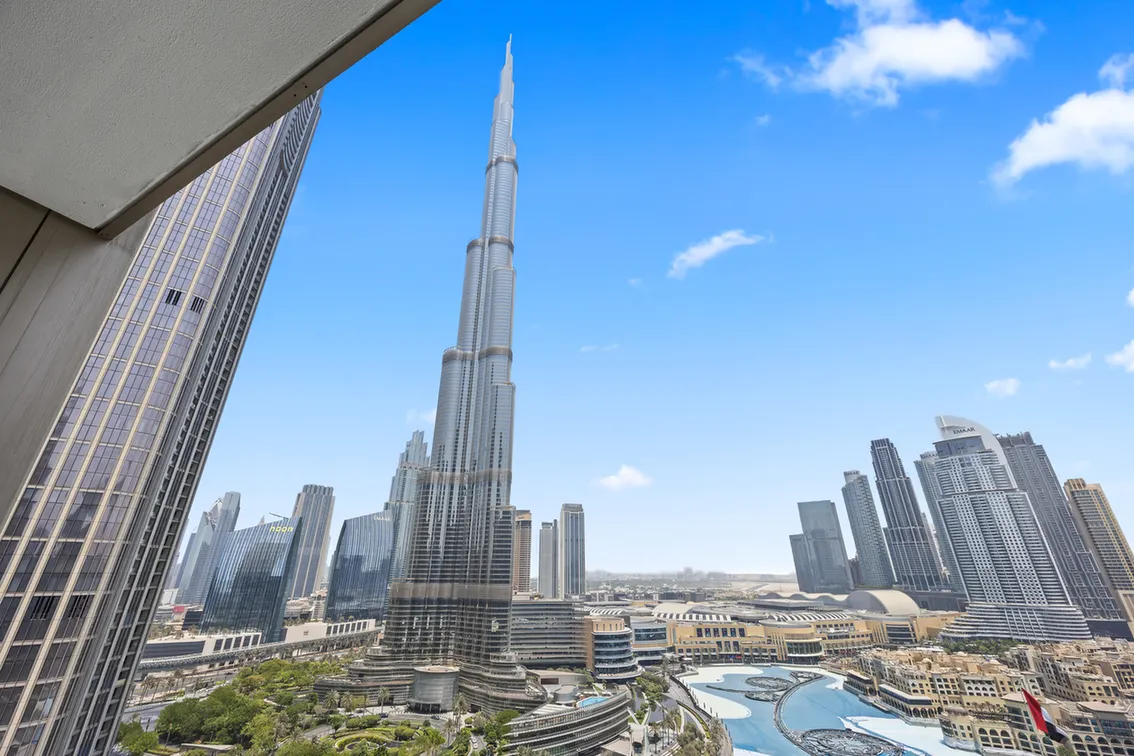 Dubai skyline with Burj Khalifa towering among glass skyscrapers under a clear blue sky, with a landscaped plaza and pools in the foreground.