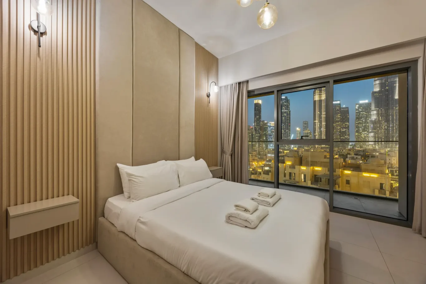 Hotel bedroom with a large window overlooking a city skyline at dusk, a white bed with folded towels on top, beige padded headboard, and wood-slatted wall panels on the left.