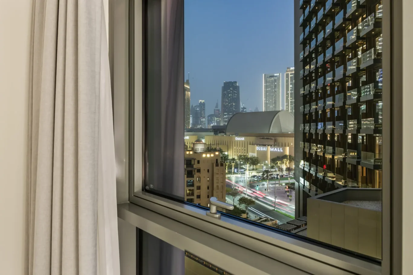 Hotel room window view of Dubai at night, with a glassy high-rise on the right and street below. Smooth curtains frame the left side.