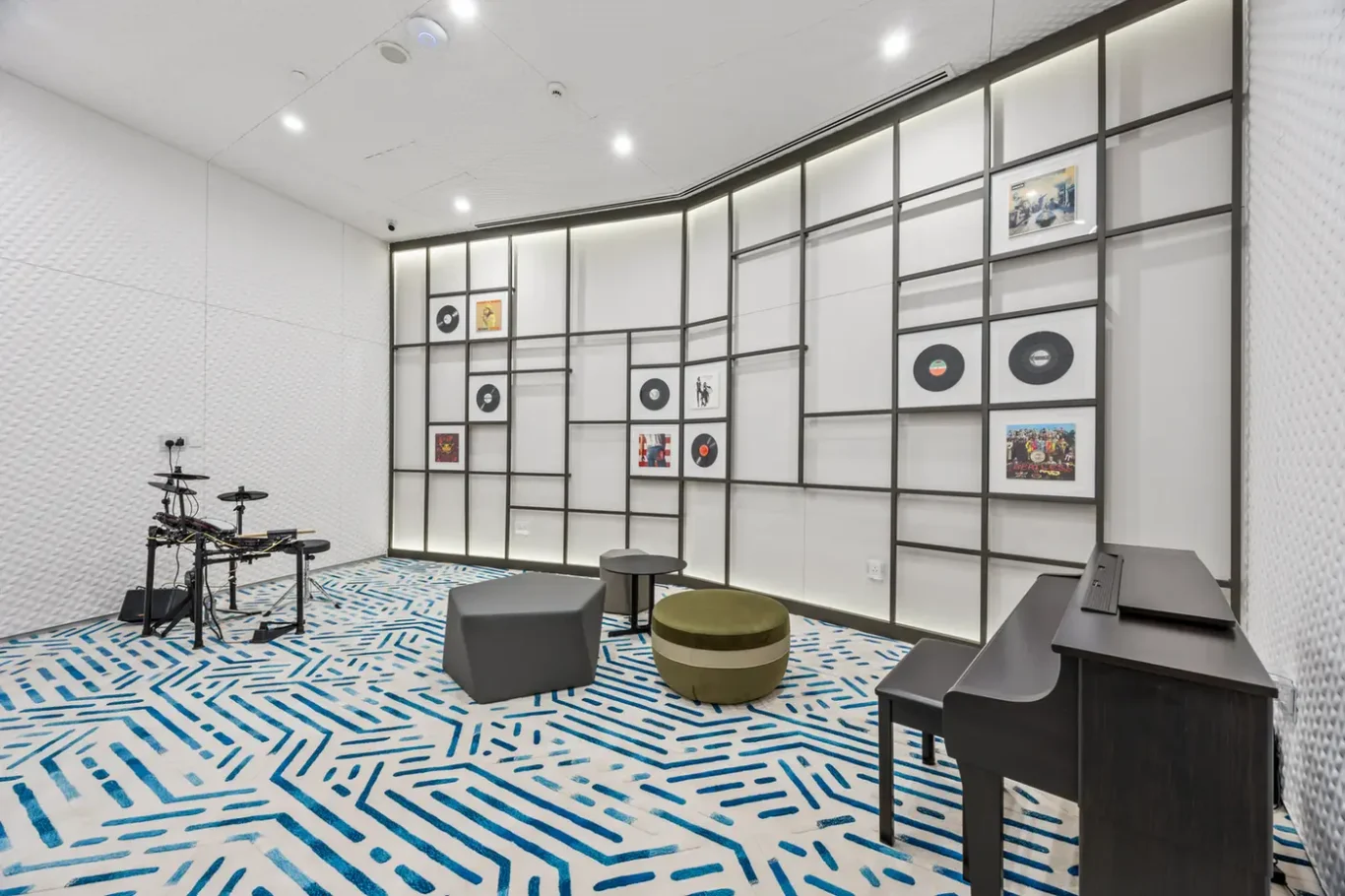 Music rehearsal room with a electronic drum kit on the left, a black piano on the right, and geometric wall panels displaying records and pictures behind.