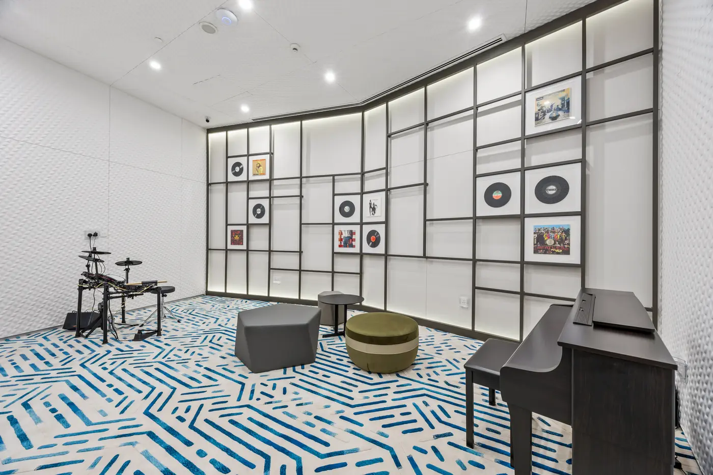 Music rehearsal room with a electronic drum kit on the left, a black piano on the right, and geometric wall panels displaying records and pictures behind.