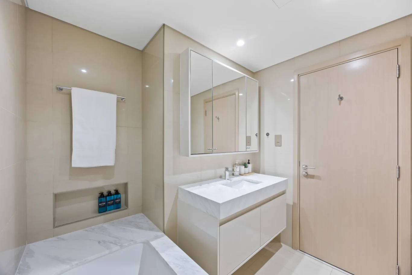 Bright contemporary bathroom with a white marble vanity, mirrored cabinet, and beige tiled walls leading to a light wood entry door.