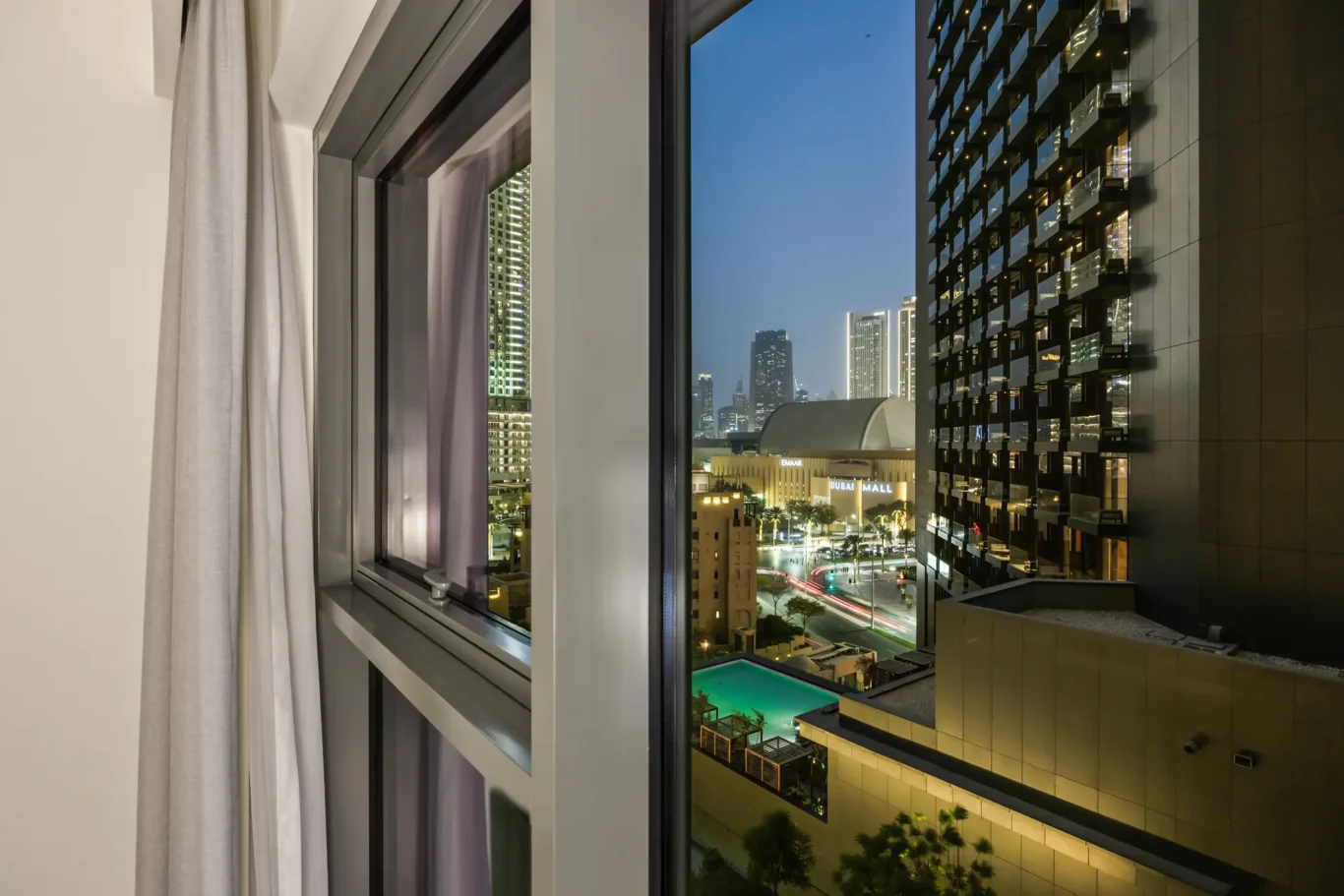 Nighttime city view from a hotel room window: illuminated skyscrapers, a glass-paneled building, and the Dubai Mall sign in the distance.