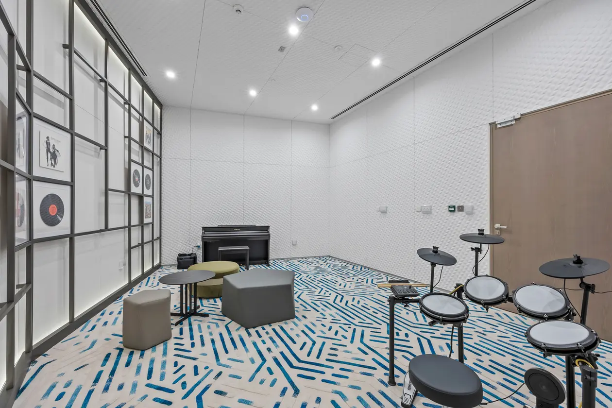 Rehearsal room with a drum set on the right, a black piano against the back wall, and modern seating on a blue geometric carpet.