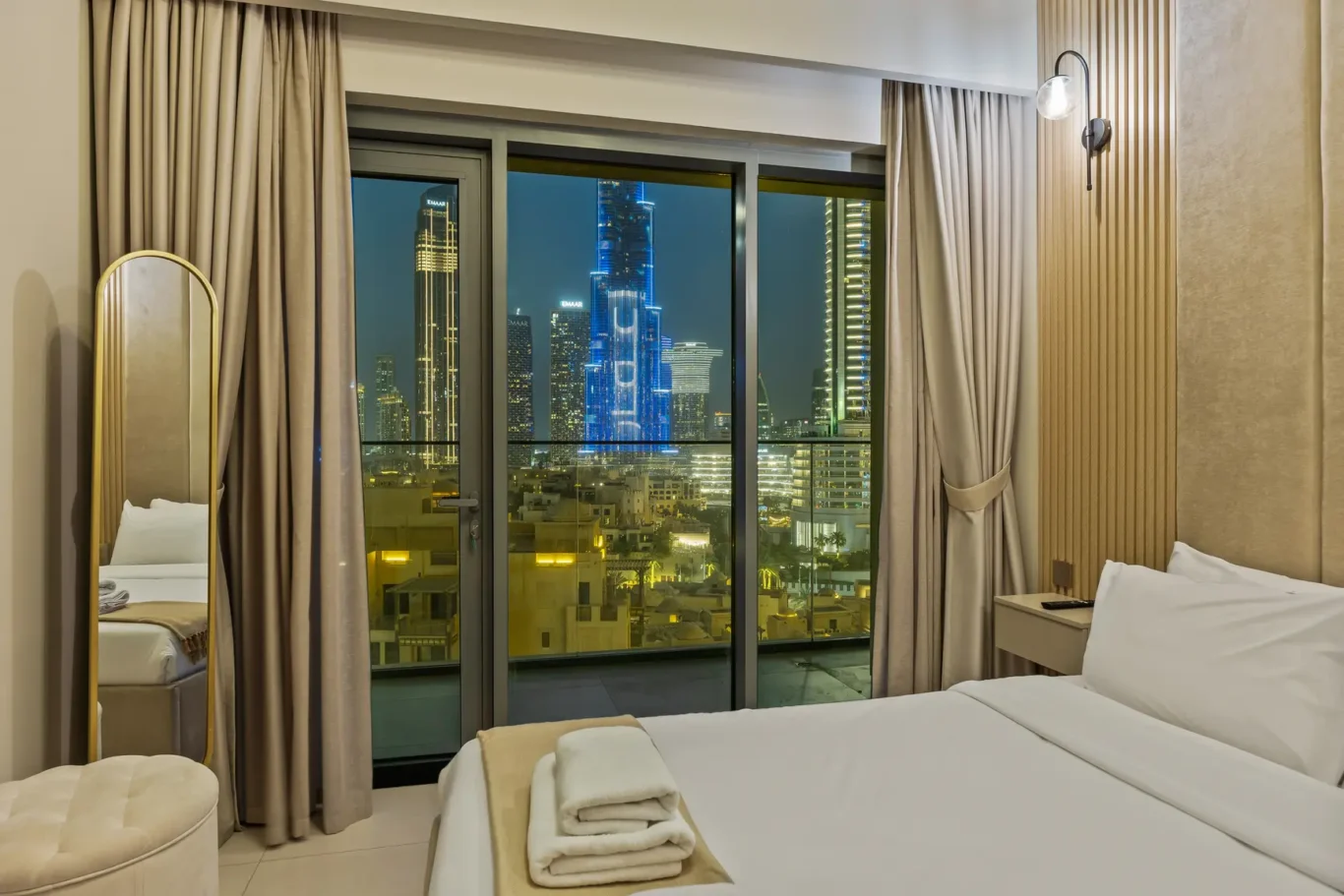Hotel room with a king bed, folded towels at the foot, and a night city skyline visible through glass balcony doors.