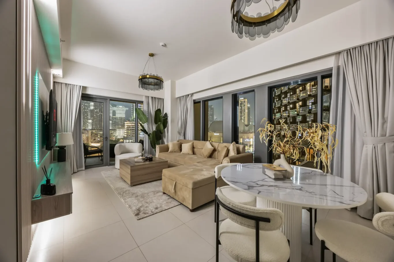 Spacious modern living room with beige sectional sofa, round marble dining table, and city skyline visible through large windows.