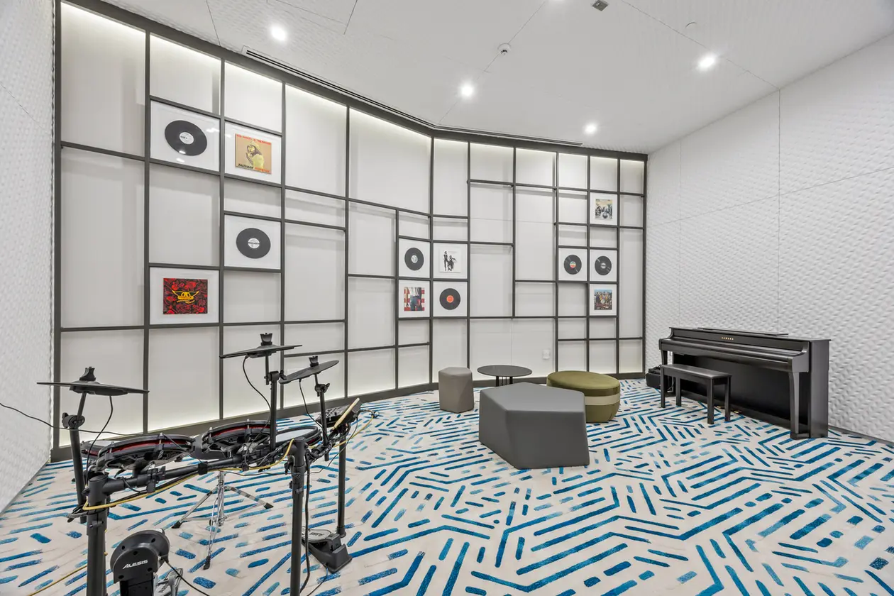 Music room with a wall grid of panels, vinyl records, and framed album covers; electronic drum kit in the foreground and a black piano on the right.