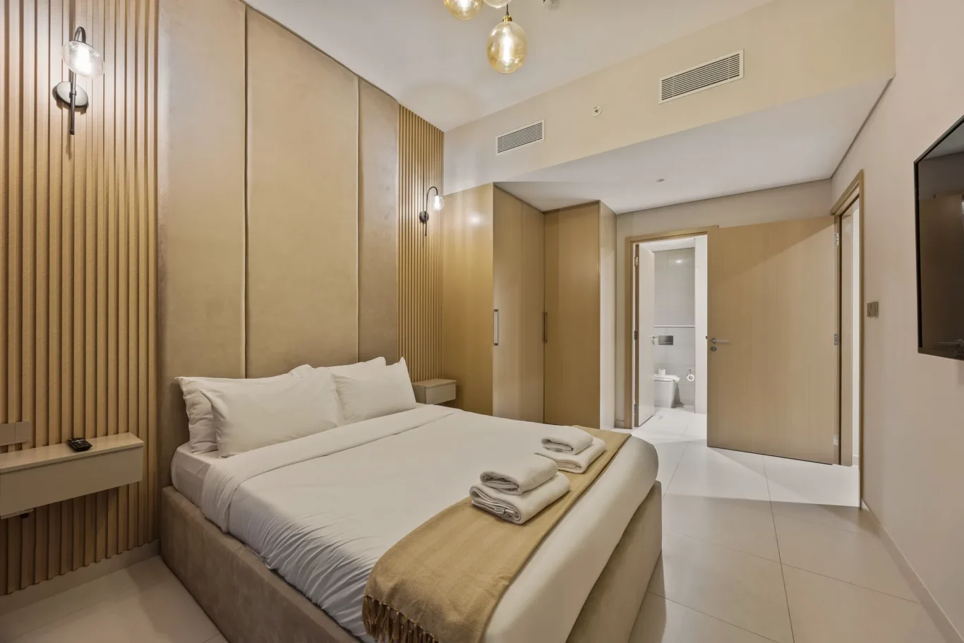 Modern hotel bedroom with a king bed, beige upholstered headboard, vertical wood paneling, and bedside tables with folded towels on the bed edge.