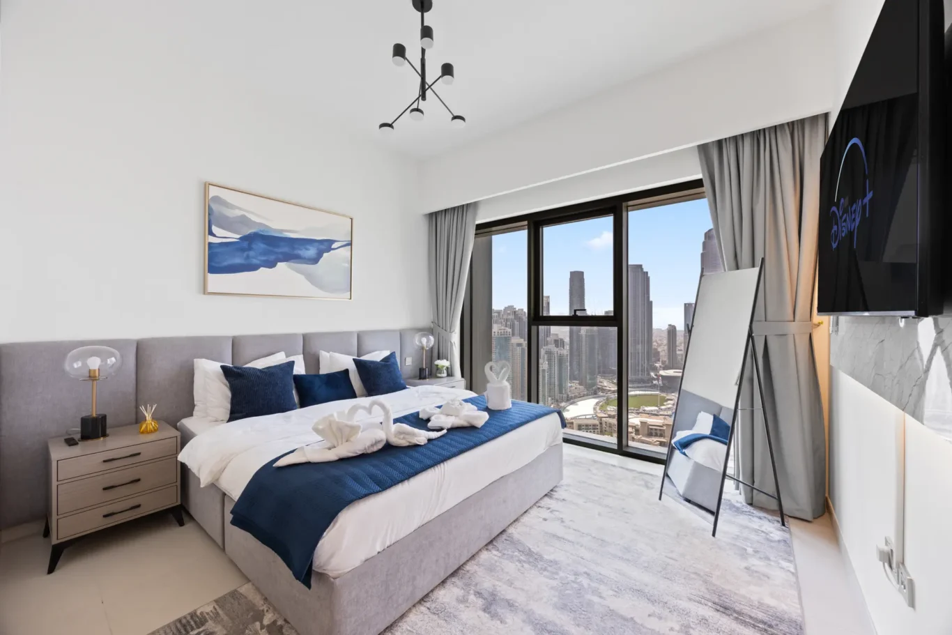 Modern hotel bedroom with a king bed, gray upholstered headboard, blue and white bedding, nightstands, and a city skyline view through large windows.