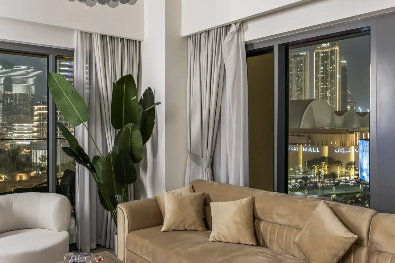 Luxurious living room at night with a beige sectional sofa, cream pillows, a tall tropical plant, and city lights seen through large windows.