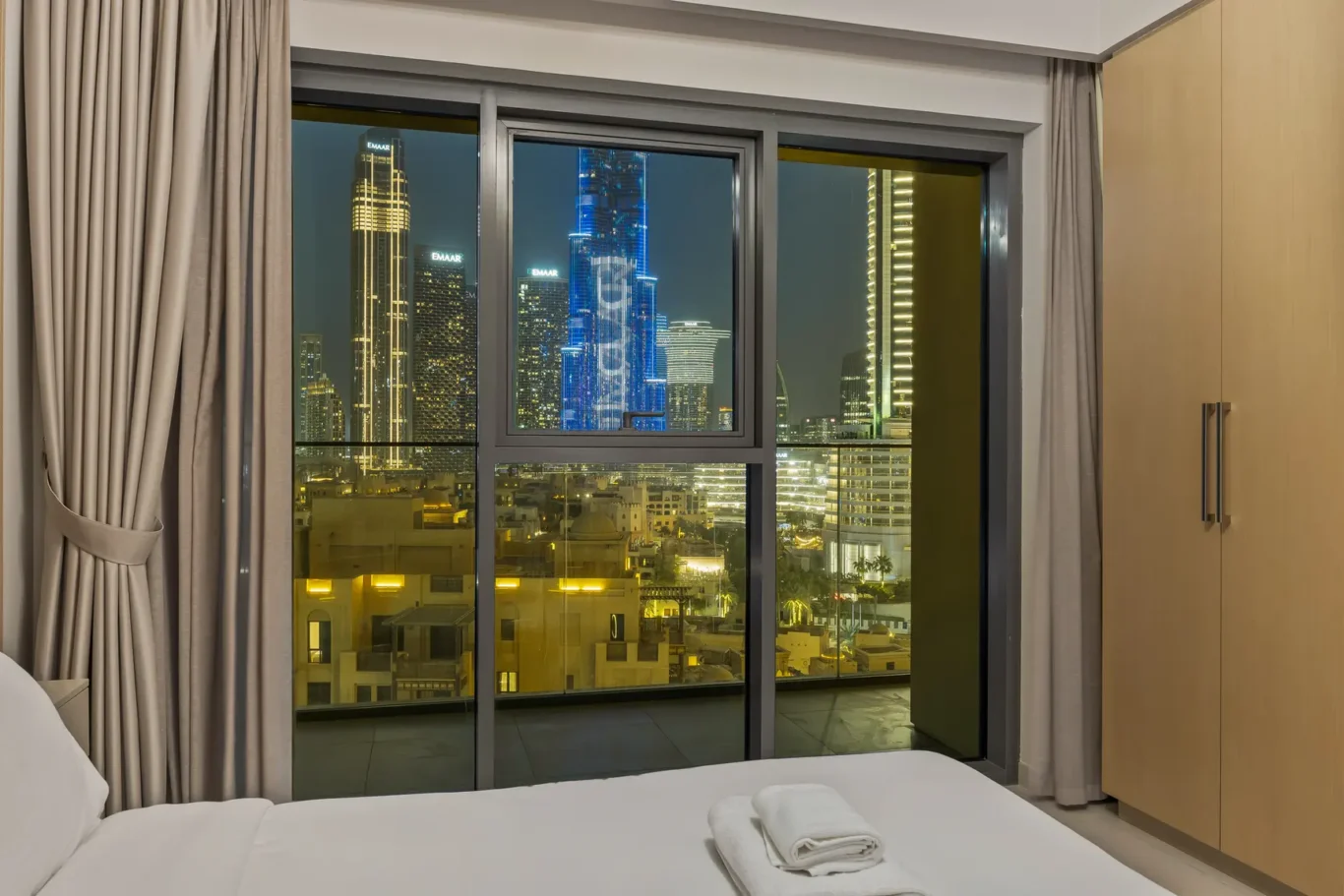 Hotel room at night with a large window showing a lit city skyline; bed in the foreground and beige curtains on both sides.